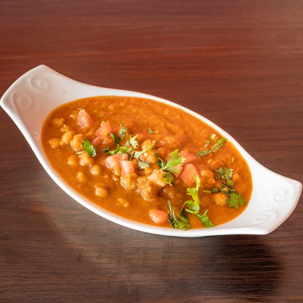 A white, ornate bowl of Indian chana masala with chickpeas, tomatoes, and cilantro on a wooden table.