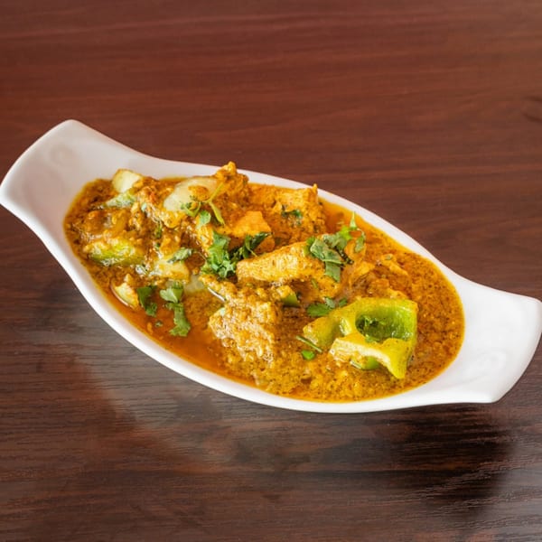 A white boat-shaped dish filled with Indian chicken curry, green peppers, and cilantro on a wooden table.
